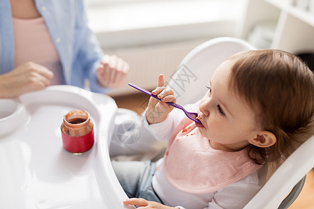 食物,孩子,喂养人的小女婴着勺子坐高椅子上,家里的厨房里吃罐子母亲的纯净物带勺子的女婴家吃罐子里的泥图片