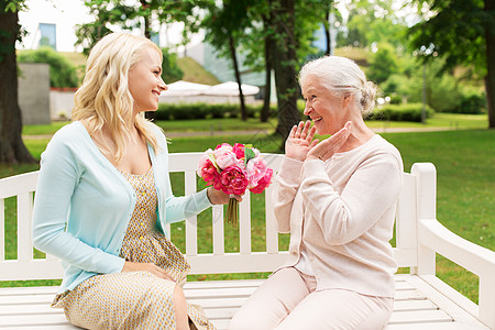 祖母绿宝石家庭,假期人的快乐的微笑的轻女儿送花给她的老母亲坐公园的长凳上女儿公园给迈的母亲送花背景