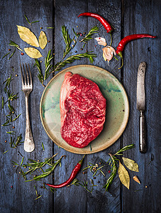 生条纹牛排用刀叉放盘子上,铺上草药香料,蓝色木制背景,顶部景色图片