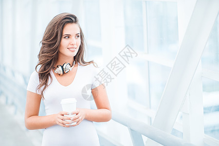 快乐的轻女人戴着音乐耳机,站桥上着咖啡杯,着城市背景向边看图片