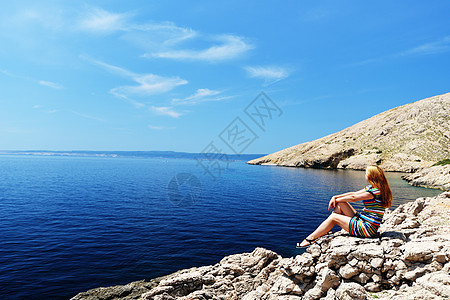美丽的轻女人坐岩石上看着大海图片