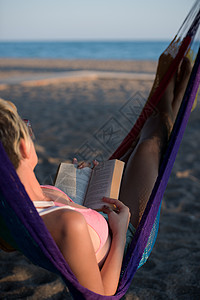 放松的女人躺沙滩上的吊床上,边看书边享受日落图片