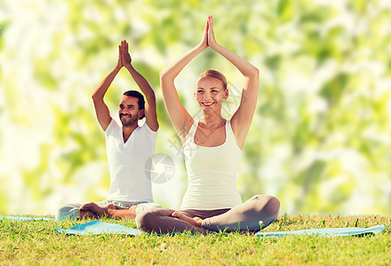 运动,健身,瑜伽人的微笑的夫妇冥想坐垫子上,举手绿色的树叶背景上图片
