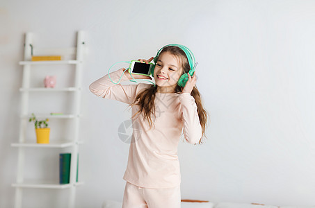 人们,孩子,睡衣派技术快乐的微笑女孩戴着耳机,用智能手机床上跳跃,家听音乐图片