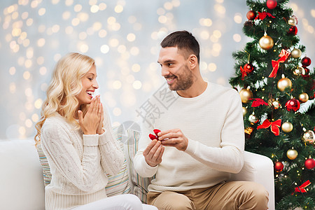 爱,夫妇,求婚人的快乐的男人小红盒子里给女人诞树灯光背景上的订婚戒指图片