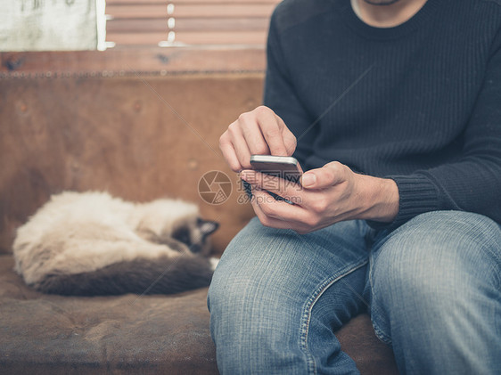 个轻人坐张旧沙发上,只猫他旁边睡觉,他正用他的智能手机图片