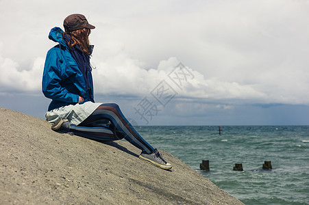 个穿着蓝色夹克的轻女人坐海边的防洪堤上背景图片