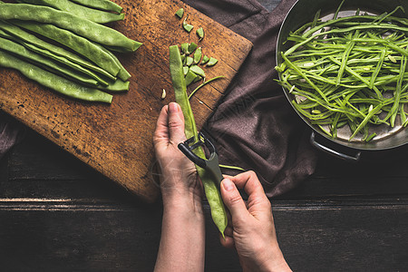 绿色法豆,黑暗厨房桌子上的准备女人的手用蔬菜剥皮机切豆切割板与绿色法豆切豆,顶部视图图片