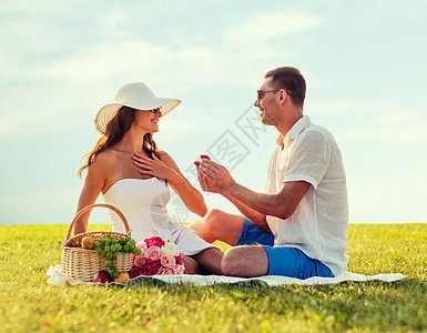 爱,约会,人,求婚假期的微笑的轻人蓝天草地背景下野餐时,把带结婚戒指的红色小礼盒送给女朋友背景图片