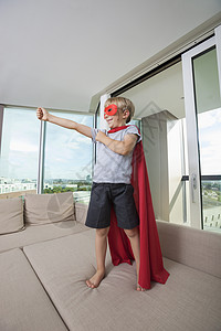 穿着超级英雄服装的微笑男孩站家里的沙发床上伸出手臂图片