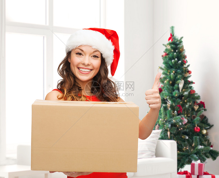 诞节,寒假,送货,手势人的微笑的女人戴着诞老人助手的帽子,包裹盒客厅里竖大拇指,背景诞树图片