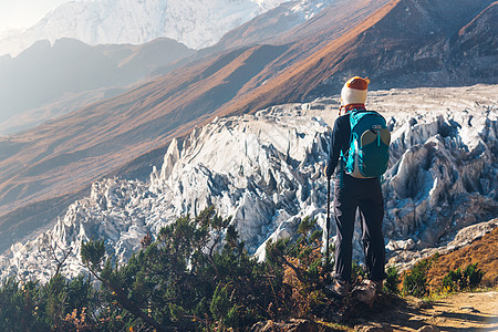 徒步山站山顶上背着背包的轻女人,日落时看着美丽的山脉冰川风景与女孩,岩石与雪峰,蓝天尼泊尔徒步旅行,旅行背景