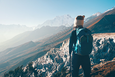 站山顶上背着背包的轻女人,日落时看着美丽的山谷风景与女孩,岩石与雪峰,山丘,蓝天尼泊尔徒步旅行,旅行老式图片