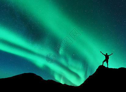 挪威山峰上站着的人的北极光轮廓,举手臂北极光快乐的人天空中星星绿色的北极光夜间景观与极光图片