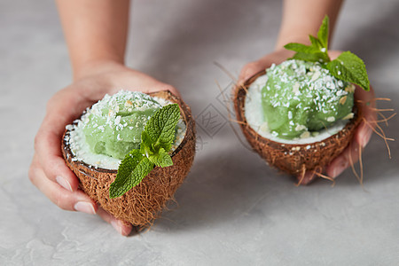 女人的手着两半椰子自制的绿色冰淇淋,灰色的混凝土背景素食者的夏季甜点个女孩着两半椰子与绿色薄荷山梨与薄荷图片