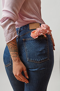漂亮的女穿着牛仔裤,背部口袋里新鲜的珊瑚色玫瑰,浅灰色背景上粉红色衬衫文字的位置女母亲节的轻漂亮的女人图片