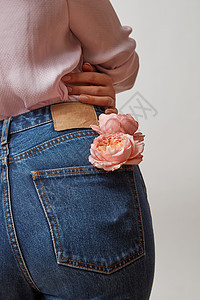 轻的女孩穿牛仔裤粉红色衬衫后与天然花玫瑰个口袋灰色的背景,女士母亲节的完美的女人底部穿着蓝色牛图片
