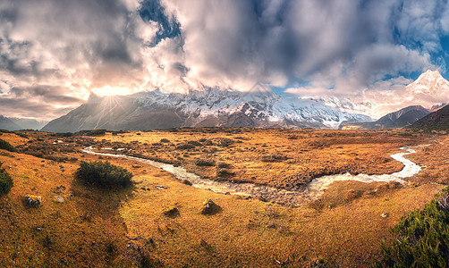 全景景观与雪覆盖的山峰云,太阳,小河,橙色的草,多云的天空五颜六色的日出五颜六色的风景与草地,小溪,雪岩尼泊尔的秋天图片