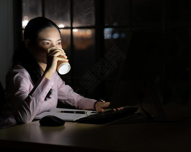 女商人深夜使用台式电脑工作时喝热咖啡图片