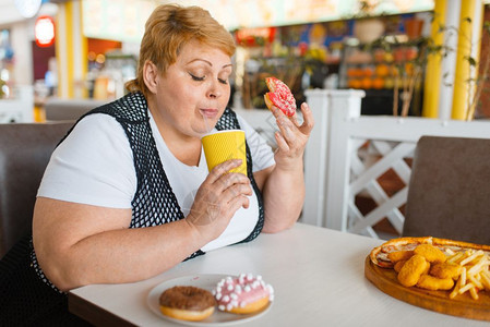 胖女人在快餐厅吃甜圈不健康的食物胖女人在餐桌上吃垃圾晚饭肥胖问题女人在快餐厅吃甜圈背景图片