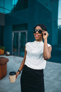 在太阳镜中快乐的女商人在室外休息时用纸板咖啡杯休息办公室建筑以背景为穿裙子和白衬衫的黑人女商在太阳镜中用咖啡休息的女商人图片