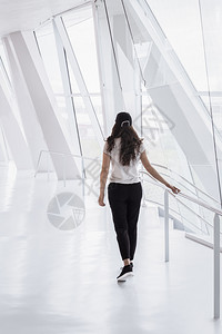 线性大楼年轻女穿着便衣走在一条明亮的白色走廊上从有高窗和现代柱子的办公大楼走出来背景