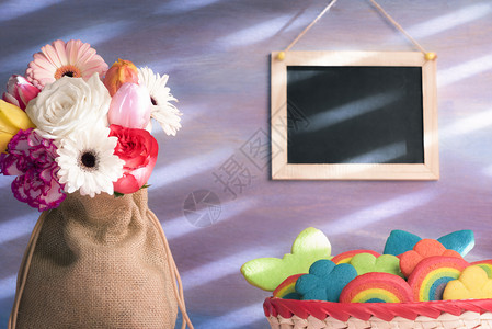 黄麻袋里花朵的美丽束一篮子上面有多种形状的彩色饼干和挂在紫墙上的空白黑板图片