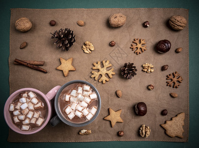 上面有两杯小棉花糖和热巧克力上面还有两杯小棉花糖和热巧克力放在布满坚果和冬糖的棕色桌布上图片