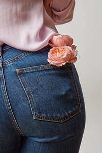 身穿牛仔裤的美丽女屁股鲜红的珊瑚玫瑰在浅灰色背景的后口袋里穿着牛仔裤和粉红色的衬衫背景图片