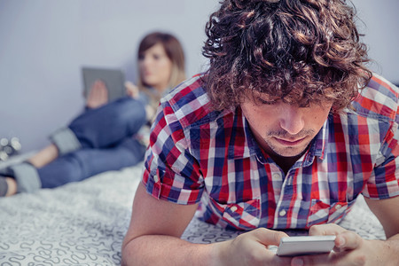 穿着智能手机的年轻英俊男子躺在床上的智能手机和美丽的女人在电子平板上阅读背景在家的休闲时间概念男人在床上使用智能手机女人在平板上举办高清图片素材