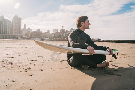 坐在沙滩上的年轻冲浪者带着他的冲浪板看海洋运动和水的概念图片