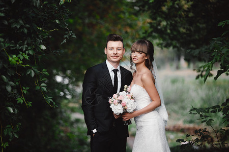 新婚夫妇郎和婚夫妇站着并拿花束盛放粉色和紫的花绿园里有色的带丝关于自然的婚礼仪式图片