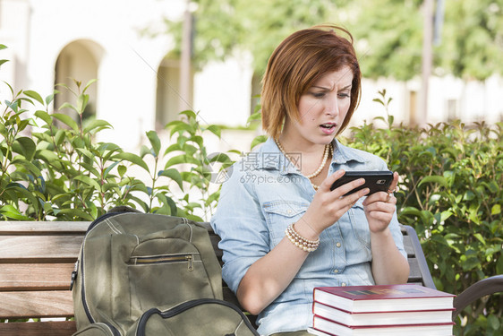 外面的年轻漂亮女学生惊呆了背着包和书本坐在长椅上手机发短信图片