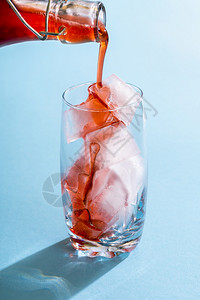 在一片装满冰块的玻璃杯中倒浆草莓糖由有机水果制成的自草莓糖浆将红水果糖浆倒在蓝底的冰块上图片