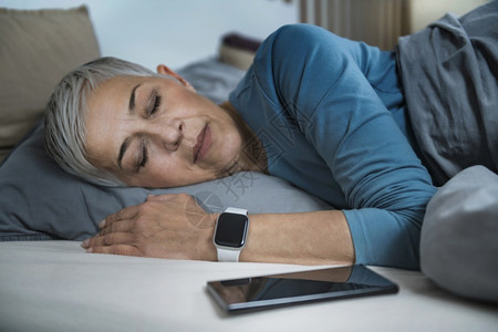 睡眠应用高科技流经的老年妇女睡在床上使用智能电话和手表来改善她的睡眠习惯图片