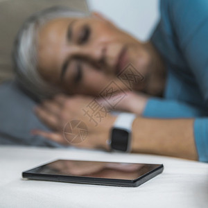 睡眠应用高科技流经的老年妇女睡在床上使用智能电话和手表来改善她的睡眠习惯图片