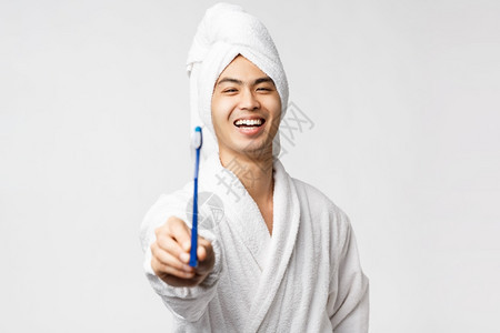 穿着浴袍和巾的年轻快乐亚洲男子展示牙刷和微笑的快乐照顾个人卫生站立白色背景美丽spa和休闲概念穿着浴袍和巾的年轻亚洲人站立白色背图片
