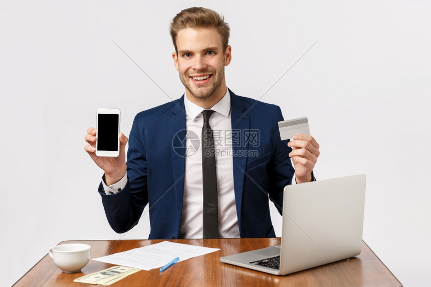 成功财富和金融概念英俊兴奋的青年商人在办公室的男创业者拿着钱笔记本和杯子的办公桌拿着信用卡显示智能手机屏幕英俊的青年创业者持有信图片