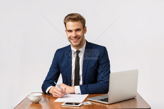 有时间赚钱英俊的办公室经理人坐在办公桌边写报告微笑回答客户拿着笔准备文件使用笔记本电脑喝杯中的咖啡赚钱英俊的办公室经理人坐在办公图片
