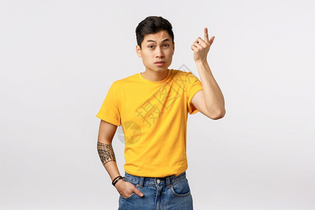 穿着黄色t恤衫的亚洲青年男子身穿黄色t恤纹身提高食用指惊慌和刺激举起眉毛如听胡言怪异对话白色背景wtf继续身穿黄色t恤衫惊慌和刺图片
