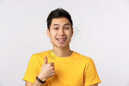 近身演播室拍摄着快乐的有吸引力穿黄色t恤衫的亚洲男展示了拇指和微笑例如给予积极的反馈建议使用公司服务批准选择白色背景近身演播室拍图片
