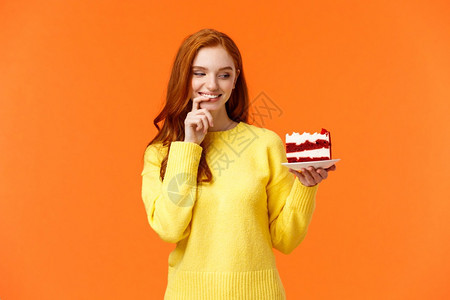 快乐和满意,的红发女人有欺骗的一天 吃美味食物,持有美味的一块蛋糕,咬嘴唇和微笑无法抗拒诱惑欲望被咬橙色背景。 快乐和满意,拿着模型高清图片素材