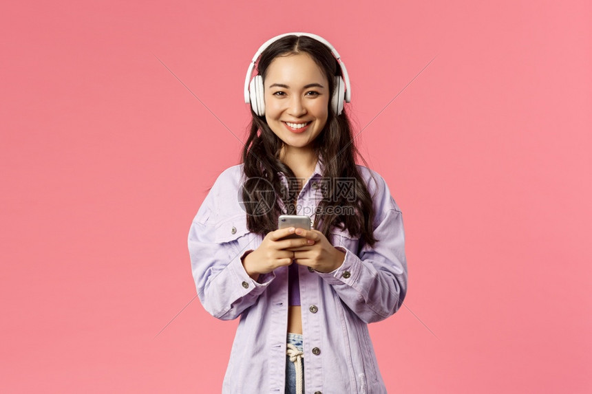 具有吸引力的现代和时尚棕褐色韩国人肖像使用手机的耳中女学生监听音乐为出去制作新的播放列表站着粉红背景具有吸引力的现代和时尚棕色韩图片