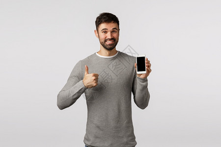 身穿灰色毛衣的英俊和快乐胡子男建议使用应程序或像新的杂货店购物场所持有智能手机展示拇指以认可图片