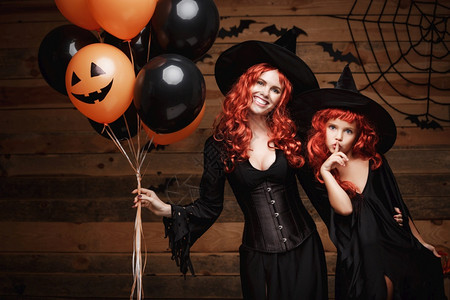 欢乐的母亲和她女儿穿着巫服装庆祝圣殿扮成橙色和黑的气球在木制工作室背景上披橙色和黑的气球蜘蛛网欢乐的母亲和她女儿穿着巫装图片