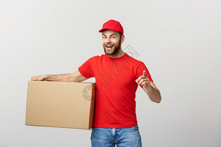 快乐的年轻英俊送货员拿着纸板盒站在灰色背景下举起拇指快乐的年轻英俊送货员拿着纸板盒图片