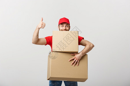 快乐的年轻英俊送货员拿着纸板盒站在灰色背景下举起拇指快乐的年轻英俊送货员拿着纸板盒图片