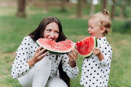 抱着西瓜两人快乐的母亲和女儿在夏季公园吃西瓜快乐的笑脸家庭在公园吃西瓜母亲和女儿一起度过时间饮食维生素健康的食物概念选择焦点饮食健康的物概念背景