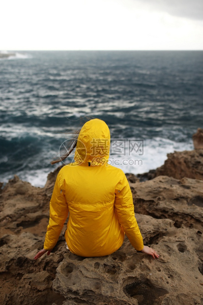 身着黄色雨衣坐在悬崖上看着海浪的大同时也享受着美丽的海景在雨天中下云雾的春季天气中岩石海滩上图片