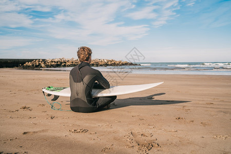 坐在沙滩上的年轻冲浪者带着他的冲浪板看海洋运动和水的概念图片